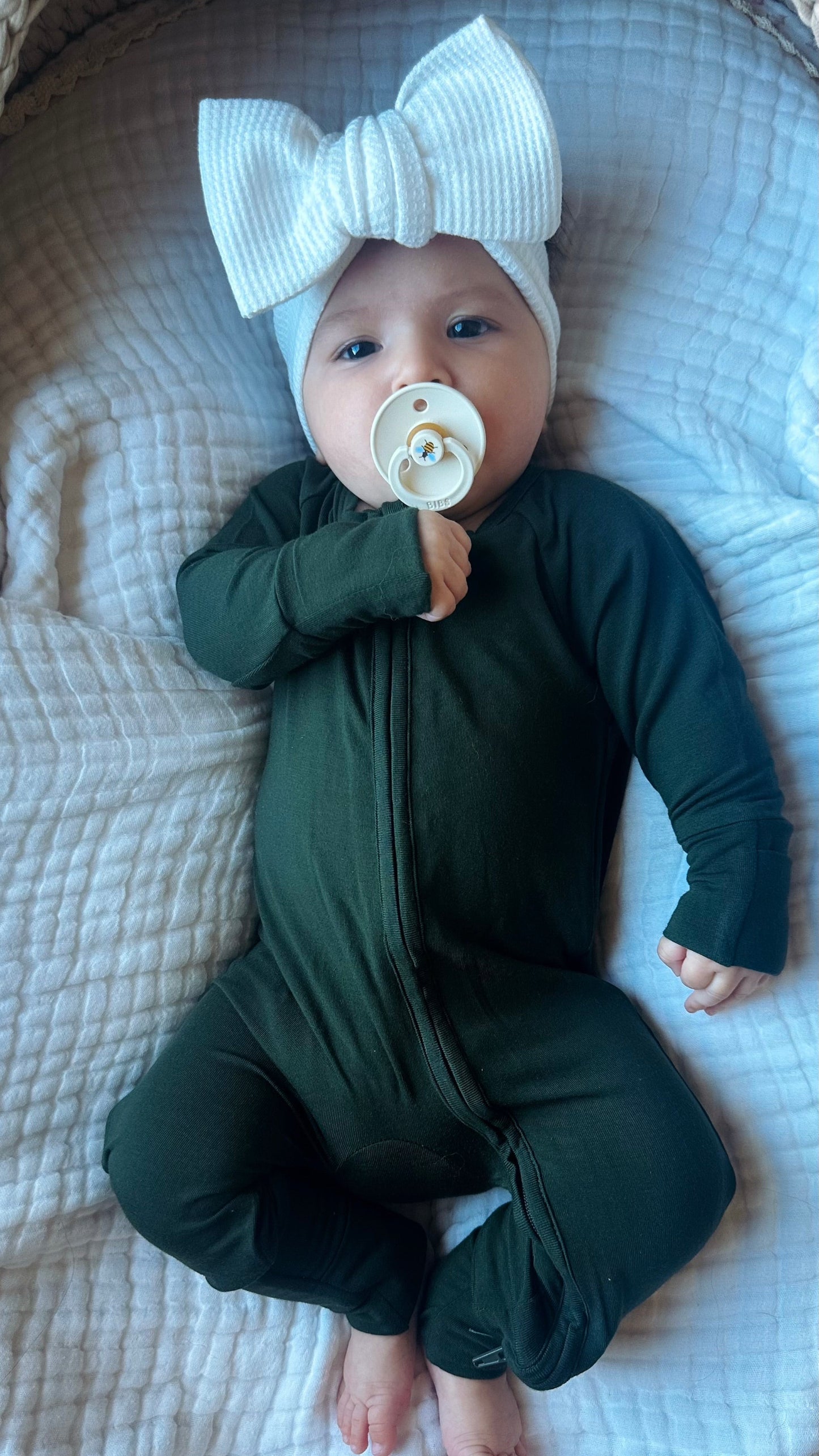 Baby in a green outfit with a white bow and pacifier, lying on a textured surface.