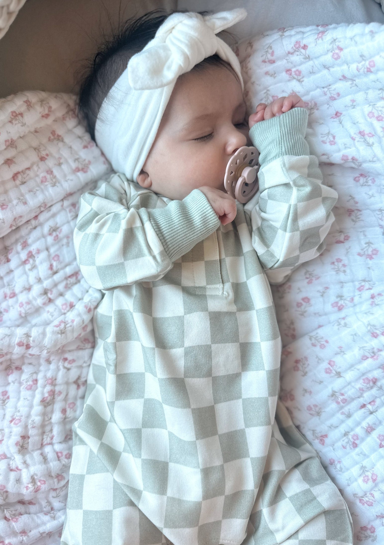 Baby in a checkered outfit with a pacifier and headband, lying on a soft surface.